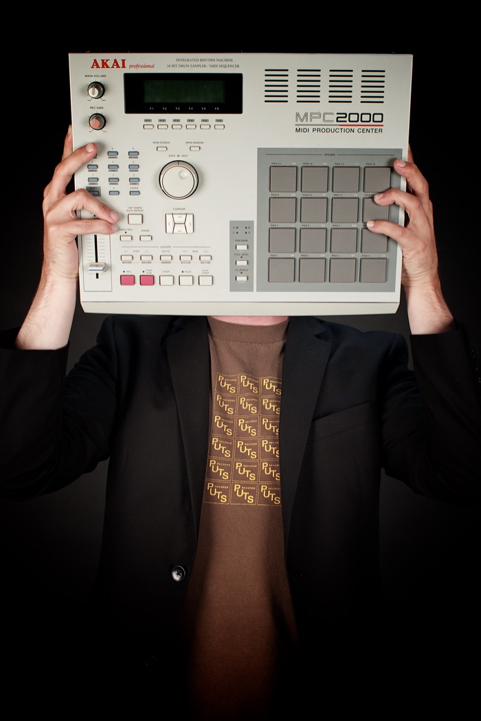Tizz Tarkington holding an Akai MPC2000 sampler in front of his face, wearing a black blazer.